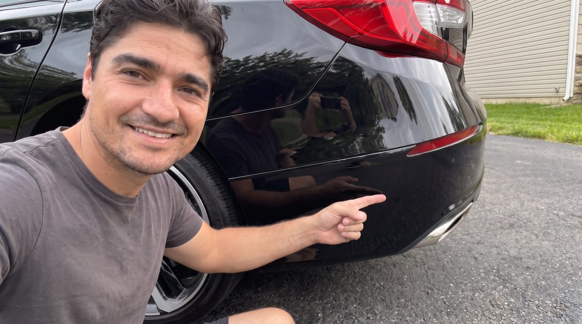 Rick kneeling beside his rear bumper, pointing at the repaired corner and smiling.
